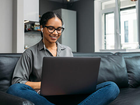 A young woman is sitting on a couch and smiling while looking at her laptop. She is wearing headphones and has her hair in a bun.