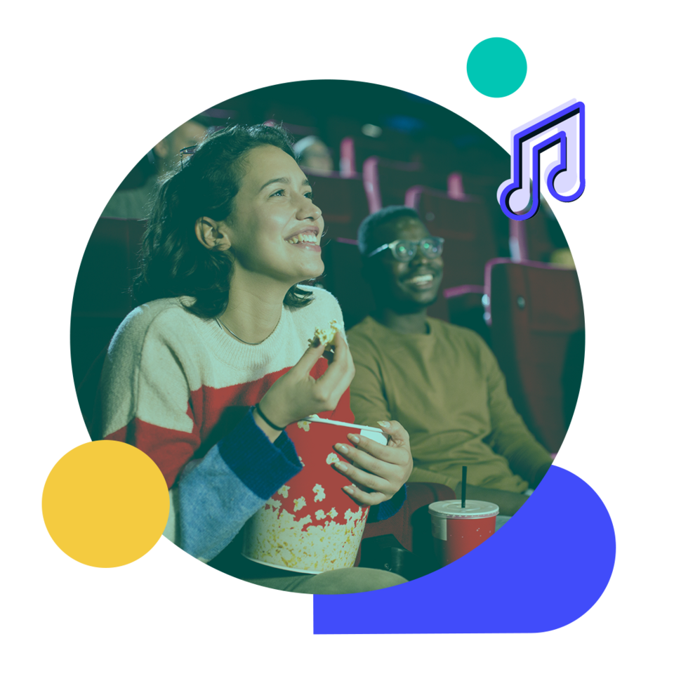 A young woman is sitting in a movie theater seat, eating popcorn and smiling. A young man is seated on her right also smiling.